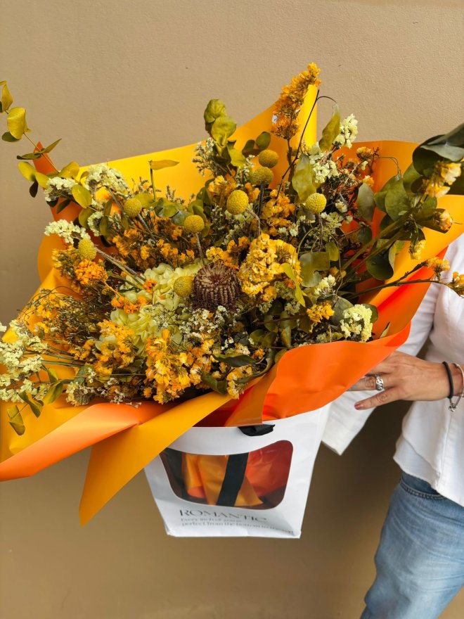 A bouquet of golden yellow dry flowers and preserved botanicals wrapped in bright orange paper and carried in a white gift bag.