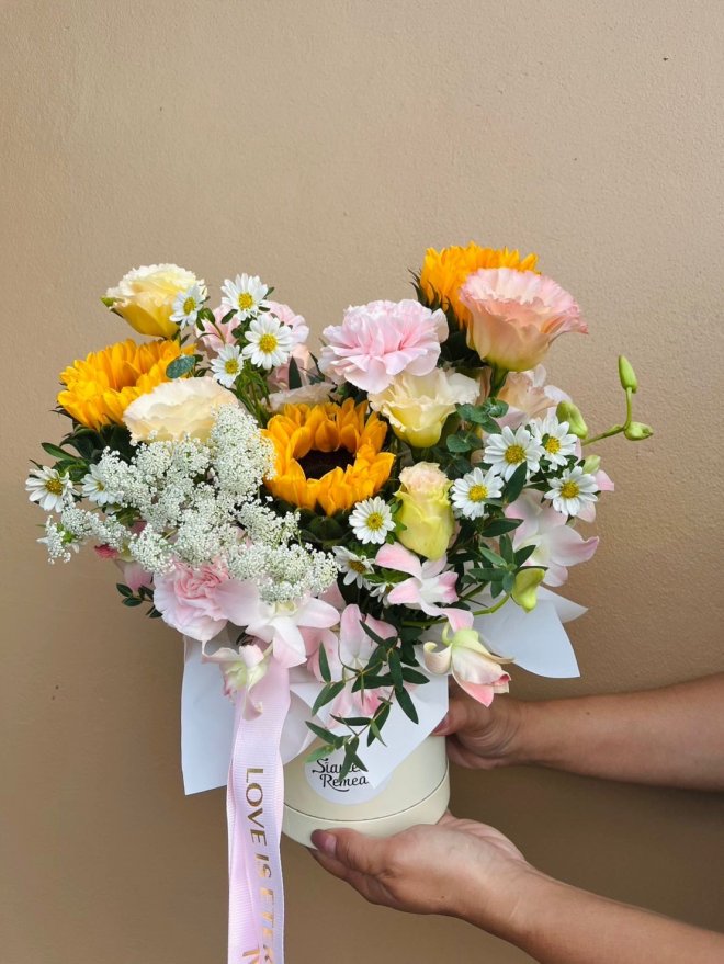 A cheerful vase bouquet with sunflowers, carnations, lisianthuses, daisies, and orchids, tied with a pink ribbon that reads “Love is Eternal.”