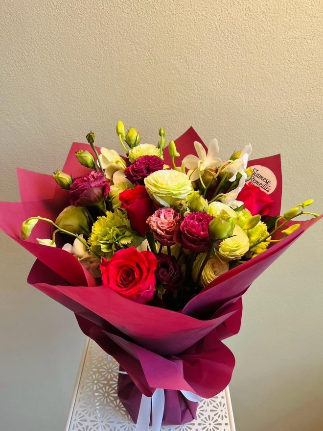 Bouquet of fuchsia roses, green carnations, lisianthus, and orchids wrapped in burgundy paper with white ribbon