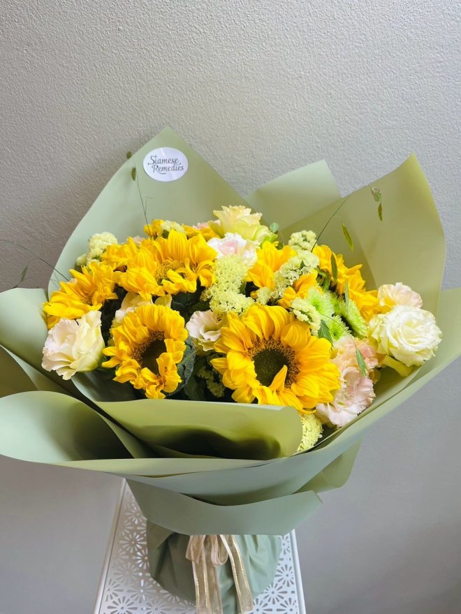 Bouquet of sunflowers, pale yellow roses, and green blooms wrapped in sage green paper.