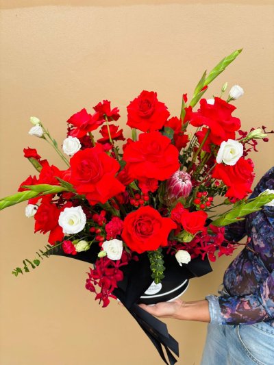 A black hat box arrangement with red roses, white lisianthuses, orchids, protea, and green accents.