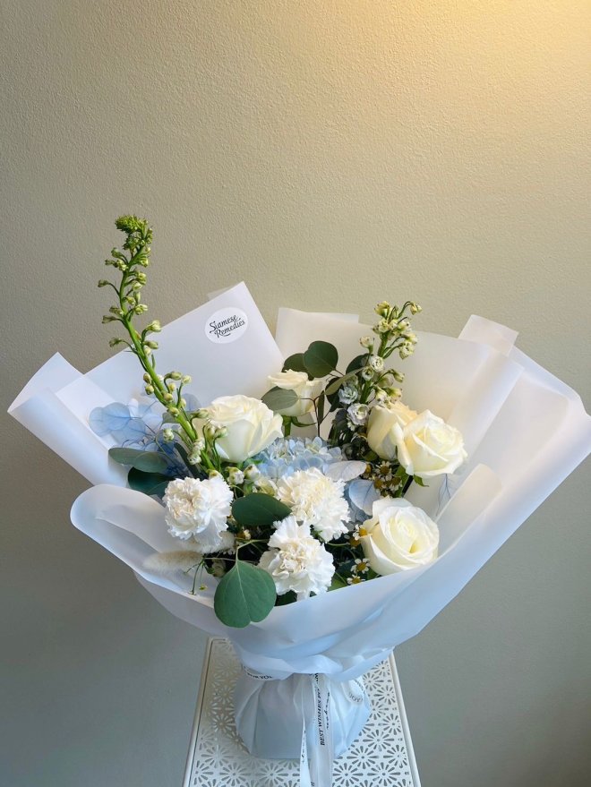 A delicate white and blue bouquet featuring ivory roses, white carnations, chamomile, and hydrangeas wrapped in frosted white paper.