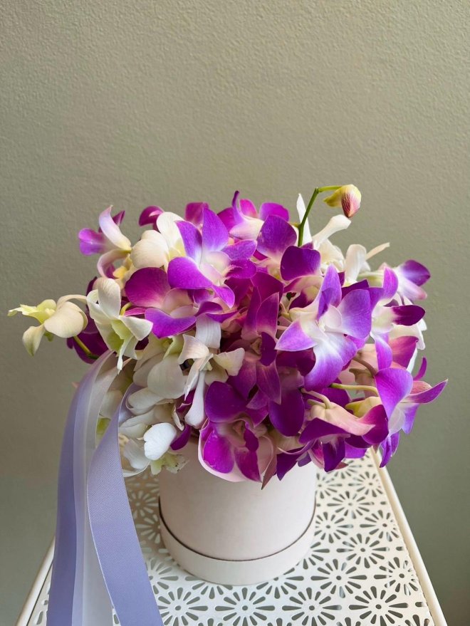 Arrangement of purple and white dendrobium orchids in a white hat box with lilac ribbon