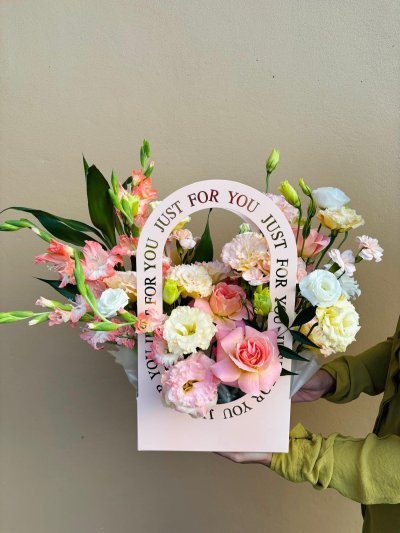 A pink floral carry bag filled with pastel roses, lisianthus, carnations, and coral gladiolus with “Just for You” printed on the handle.