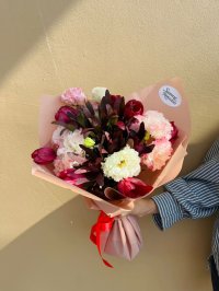 A bouquet of red tulips, burgundy leaves, and pink and cream carnations wrapped in blush paper with a red ribbon.
