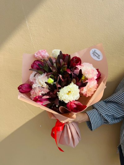 A bouquet of red tulips, burgundy leaves, and pink and cream carnations wrapped in blush paper with a red ribbon.