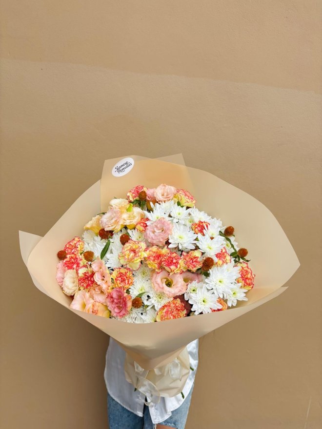 A bouquet of peach lisianthus, coral carnations, and white chrysanthemums wrapped in soft beige paper.