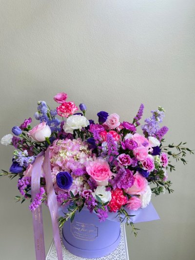 A pastel purple hatbox bouquet with pink roses, purple lisianthuses, hydrangeas, carnations, and statice, accented with lavender ribbon.