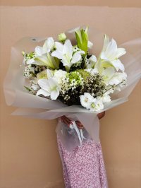 Bouquet of white lilies, lisianthuses, and wax flowers with soft green accents, wrapped in frosted paper.