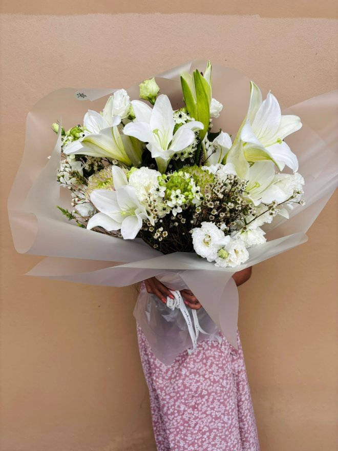 Bouquet of white lilies, lisianthuses, and wax flowers with soft green accents, wrapped in frosted paper.