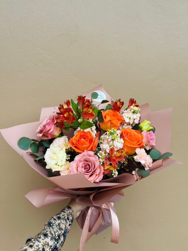A bouquet featuring orange and blush roses, lisianthuses, and red alstroemerias wrapped in dusty pink paper.