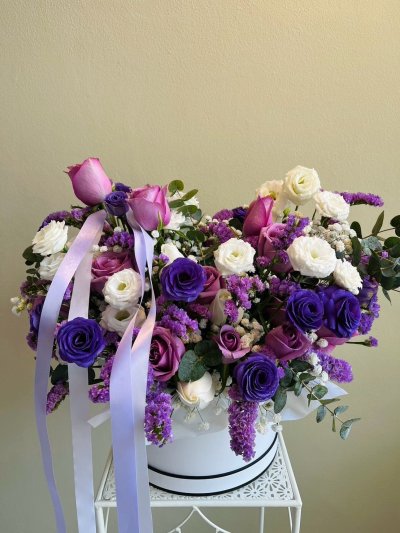 Purple and white floral arrangement with roses, lisianthus, and statice in a white hatbox with lavender satin ribbons