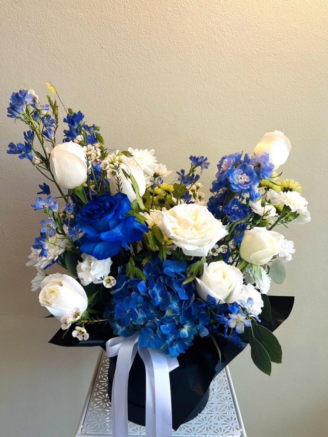 Floral arrangement of royal blue hydrangeas, white tulips, ivory roses, and blue delphiniums in a black hatbox with white ribbon