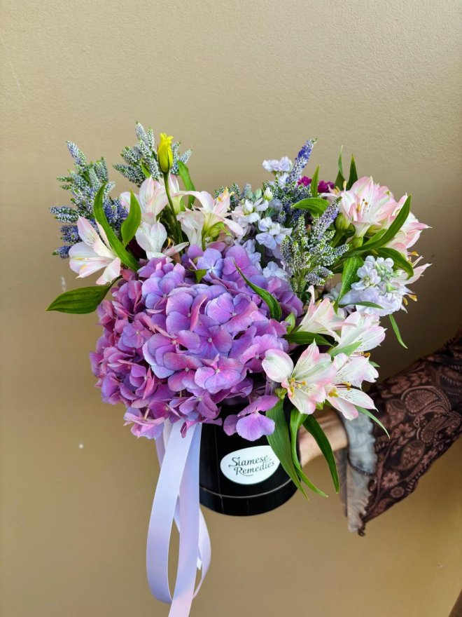 A black hat box arrangement featuring purple hydrangeas, lilac stock, pink alstroemeria, and veronica, tied with a soft lilac ribbon.