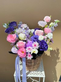 A wicker basket filled with lisianthuses, roses, carnations, and dry flowers in pink, white, and violet tones with lavender ribbon.