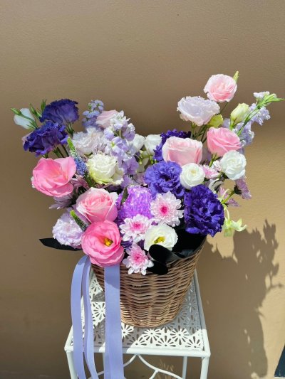 A wicker basket filled with lisianthuses, roses, carnations, and dry flowers in pink, white, and violet tones with lavender ribbon.