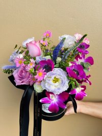 Fuchsia anemones, violet orchids, lavender lisianthus, soft pink roses, and daisies arranged in an elegant black hat box with luxe black ribbons.