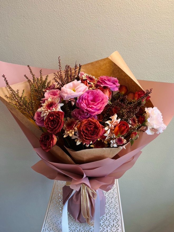 Rustic bouquet with burgundy, terracotta, and blush roses, pink lisianthus, rust mums, and wild heather in brown and pink wrap.