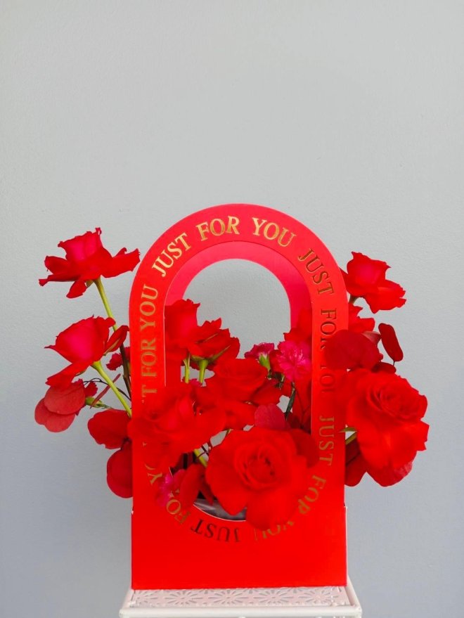 A dramatic red floral box with red roses and carnations arranged inside a “Just for You” red arch box.