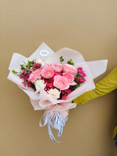 A blush-wrapped bouquet featuring pink roses, carnations, and wax flowers.