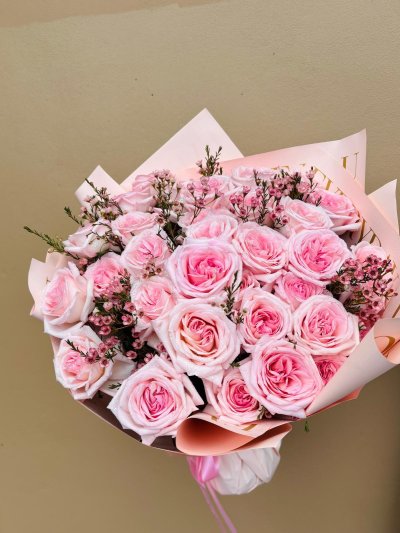 A bouquet of soft pink roses and pink wax flowers with pine-like greenery wrapped in blush paper.