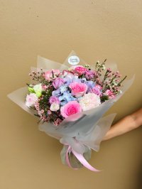 A pastel bouquet of pink roses, lavender lisianthus, hydrangeas, carnations, and wax flowers wrapped in frosted white paper.