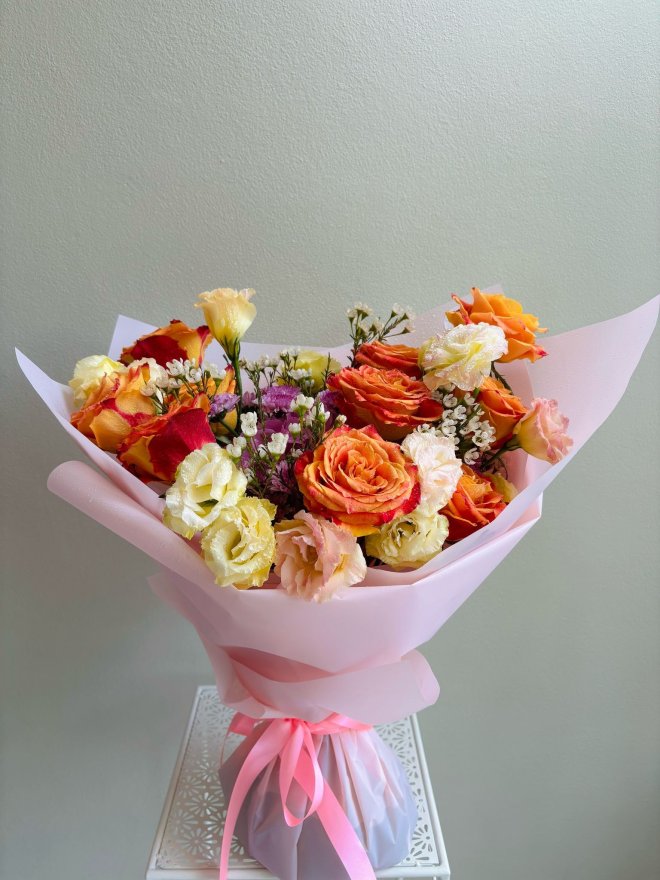 Bouquet of orange-red roses, yellow and peach lisianthus, and white filler flowers wrapped in light pink paper.