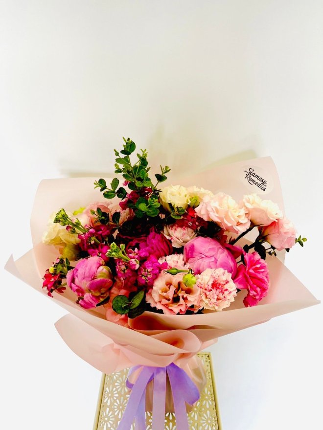 A vivid pink bouquet with peonies, carnations, lisianthuses, and roses wrapped in soft blush paper with a lavender ribbon.