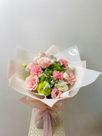 A pastel bouquet featuring pink roses, green hydrangeas, lisianthuses, and carnations wrapped in soft blush paper.