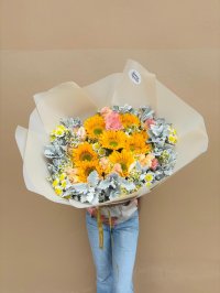 A bouquet of sunflowers, pastel carnations, and white blooms with silvery foliage, wrapped in cream paper.