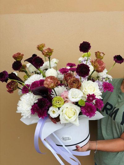 A white hat box arrangement with white roses, burgundy scabiosa, antique carnations, and purple chrysanthemums accented by calla lilies.