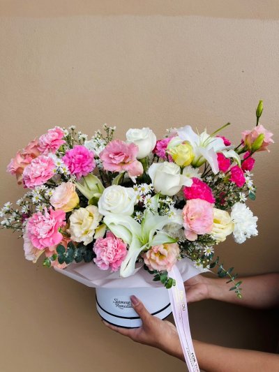 A vibrant hatbox bouquet with white roses, lilies, pink carnations, lisianthuses, and chamomile-like flowers.