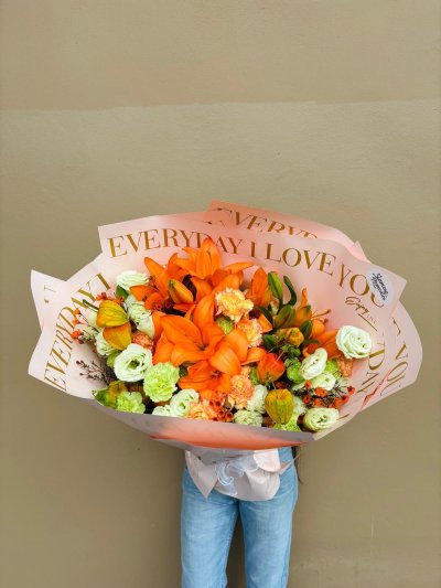 A bouquet of orange lilies, carnations, and green lisianthus wrapped in decorative “Everyday I Love You” paper.