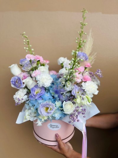 Hat box arrangement features pale lavender lisianthus, baby blue hydrangeas, delicate white roses, soft pink carnations, and airy sprigs of statice.