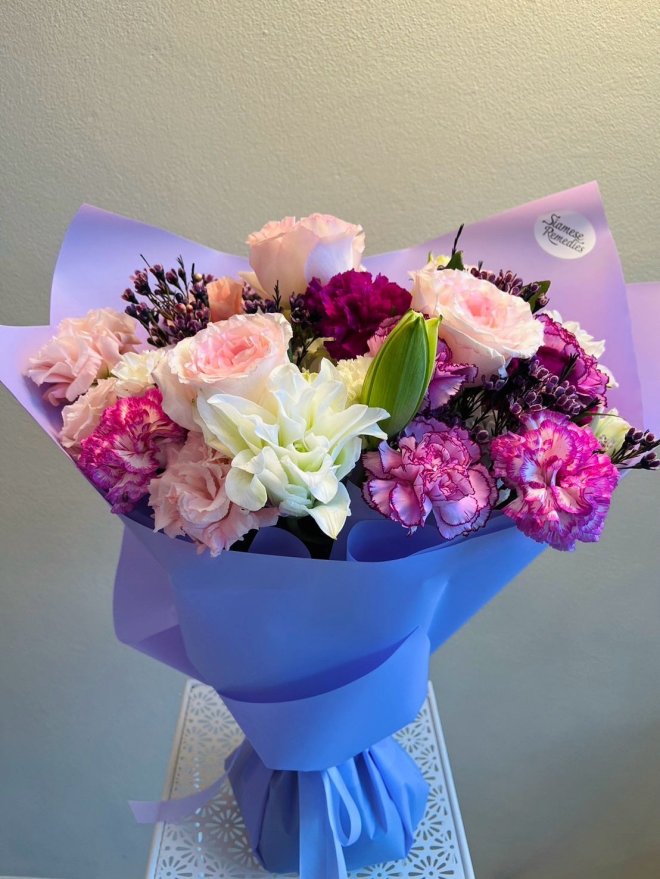 Elegant bouquet with blush roses, white lilies, violet carnations, and dark purple accents, wrapped in lavender paper.