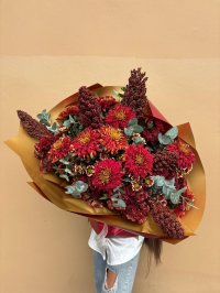 A bouquet of deep red chrysanthemums and textured blooms with eucalyptus, wrapped in golden paper.
