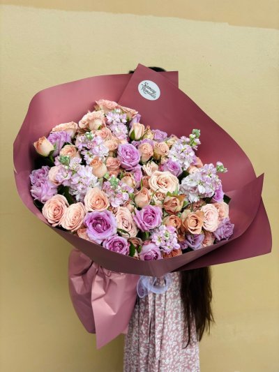 Large bouquet of peach and lavender roses with lilac stock flowers, wrapped in deep mauve paper.