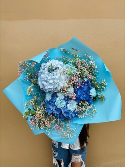 A bouquet of blue hydrangeas, blue carnations, and rainbow baby’s breath wrapped in aqua paper.