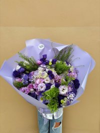 A bouquet of purple and lilac lisianthus, roses, and hydrangeas with greenery, wrapped in lavender paper.