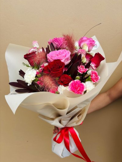 A bouquet with red roses, pink roses, carnations, and white flowers wrapped in cream paper with a red ribbon.