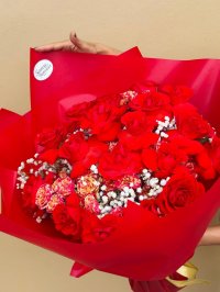 A bouquet of red roses, red-orange carnations, and white baby’s breath wrapped in bold red paper with a gold ribbon.