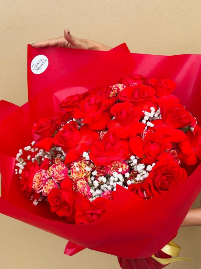A bouquet of red roses, red-orange carnations, and white baby’s breath wrapped in bold red paper with a gold ribbon.