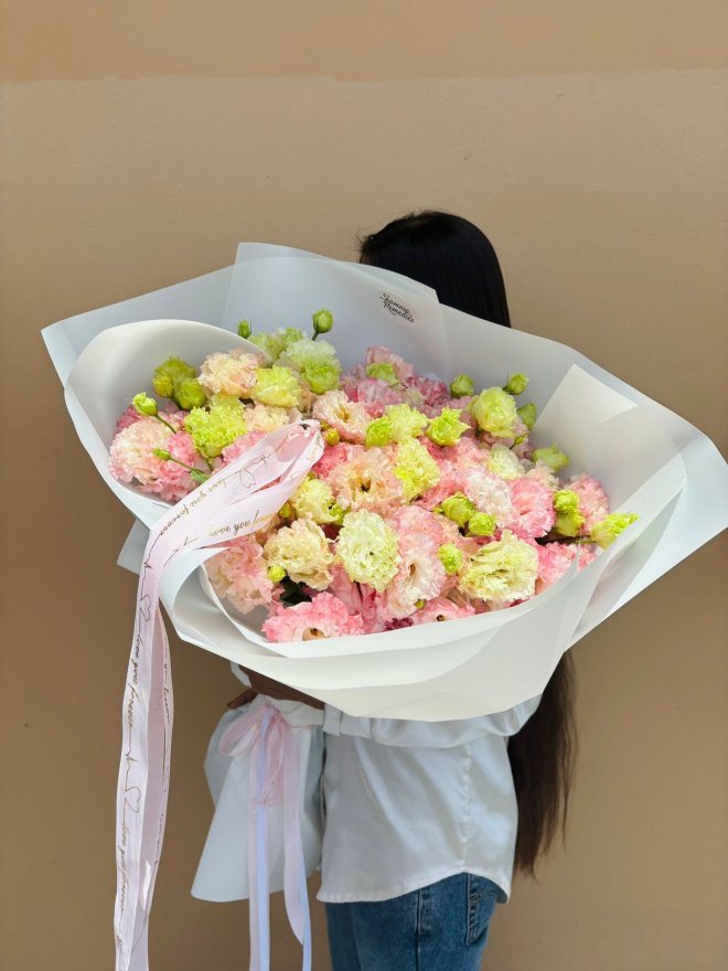A large pastel bouquet of pink, cream, and lime green carnations, wrapped in white paper with a soft pink ribbon.