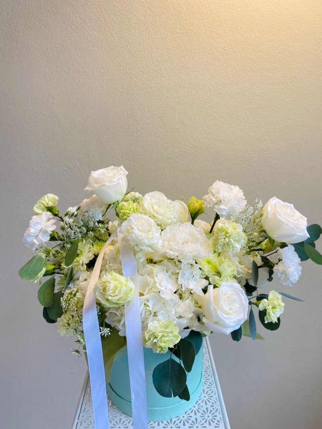 A pastel box arrangement of white roses, hydrangeas, and green-tinted carnations with soft foliage.