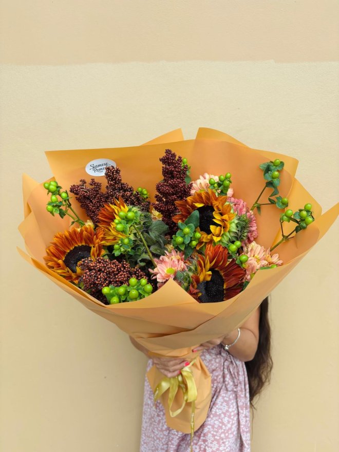 A rustic bouquet of sunflowers, red millet, chrysanthemums, and green berries wrapped in amber paper.
