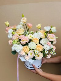 A soft peach and cream bouquet in a white hatbox featuring roses, lisianthuses, carnations, and chamomile blooms.