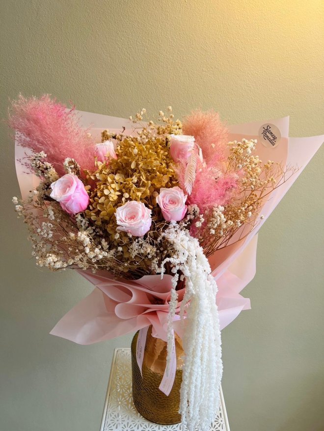 A dried floral bouquet with pink roses, golden hydrangeas, baby’s breath, and pampas in soft pink wrapping.