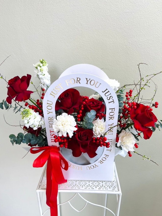 A designer floral arrangement with red roses, white carnations, red berries, and eucalyptus in a white arch frame with a red ribbon.