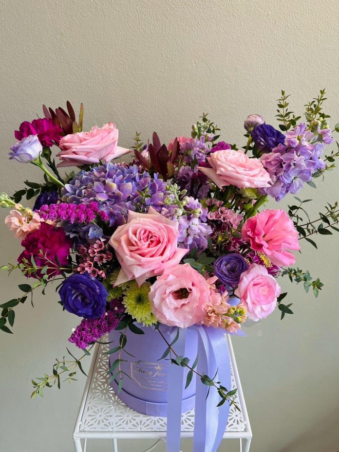 Arrangement of pink roses, purple lisianthus, lavender hydrangeas, carnations, and greenery in a lavender hatbox with lilac ribbons