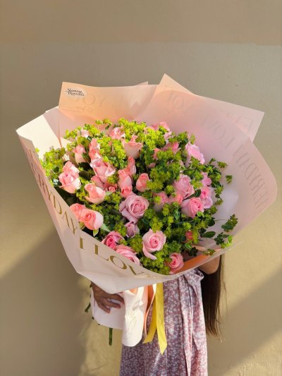 A lush bouquet of soft pink roses and vibrant green fillers wrapped in blush pink paper with a golden ribbon.